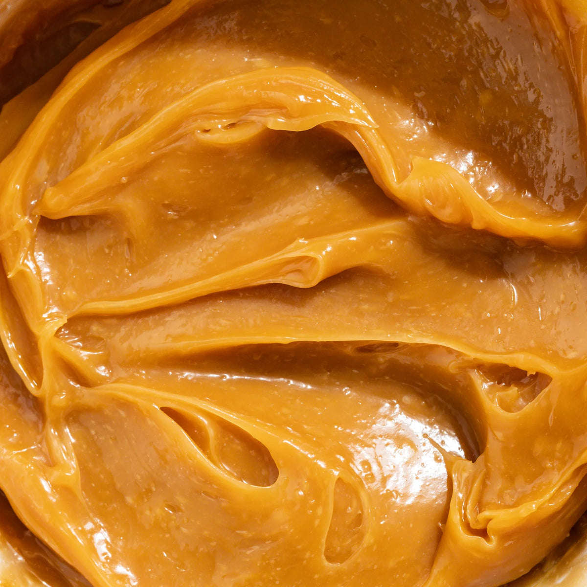 A close-up texture shot of smooth, glossy caramel-colored peanut butter with visible swirls and creamy highlights.