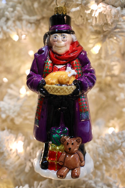 A Christmas ornament depicting a man in a purple coat and red scarf holding a basket of turkeys, standing next to a small brown teddy bear with wrapped gifts, set against a blurred white Christmas tree with warm lights.
