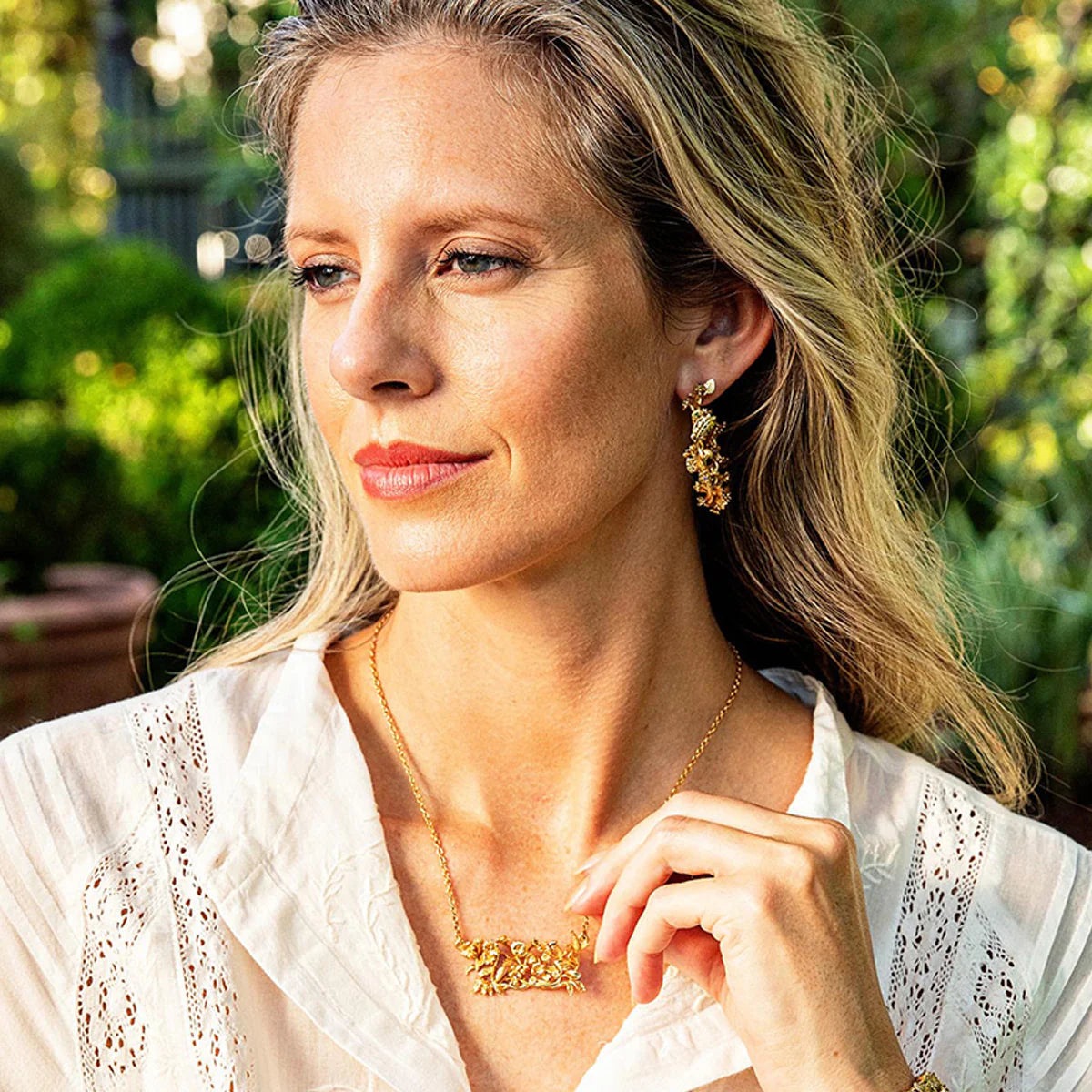 Woman wearing gold jewelry outdoors with greenery in the background