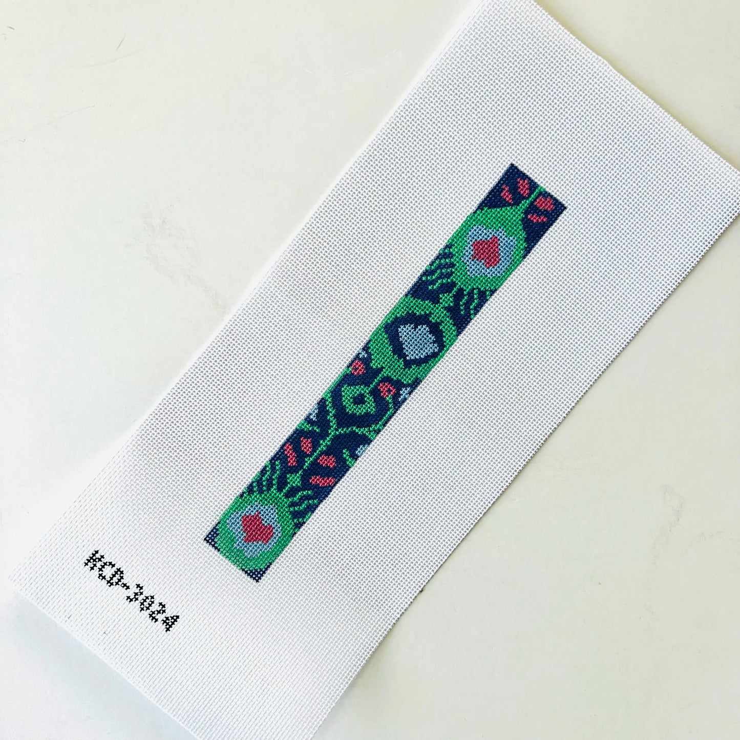 A close-up of a white woven fabric strap or band with a narrow central embroidered geometric pattern in green, blue, pink, and red, photographed on a light neutral background. Small printed text 'HCD-3024' appears near the lower left.