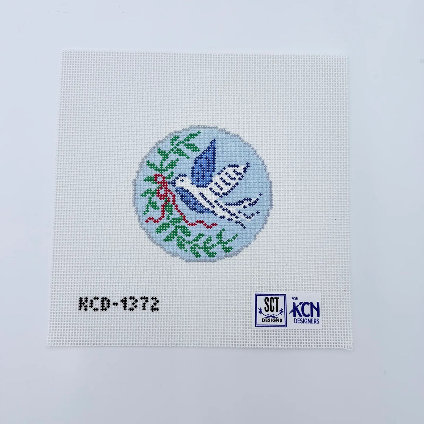Cross-stitched design of a dove with an olive branch on fabric with 'KCN' branding.