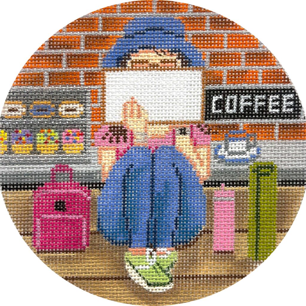 Circular needlepoint canvas with a person sitting at a coffee shop counter, surrounded by colorful elements.