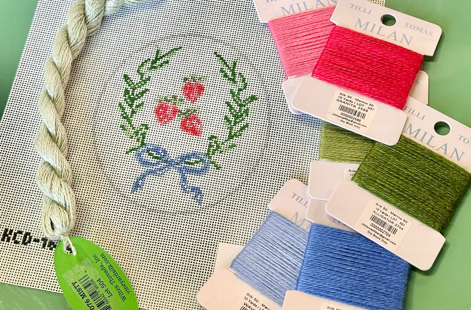 Needlepoint canvas with strawberry design and colorful thread packages on a green surface