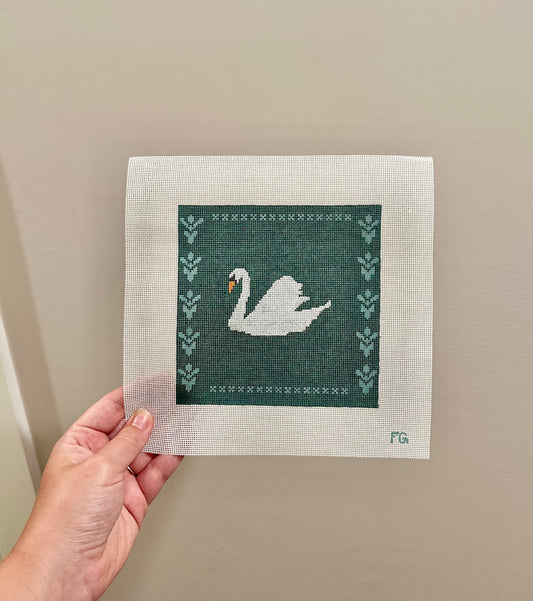 A hand holding a square needlepoint canvas featuring a white swan on a dark green background with decorative borders, set against a plain beige wall.