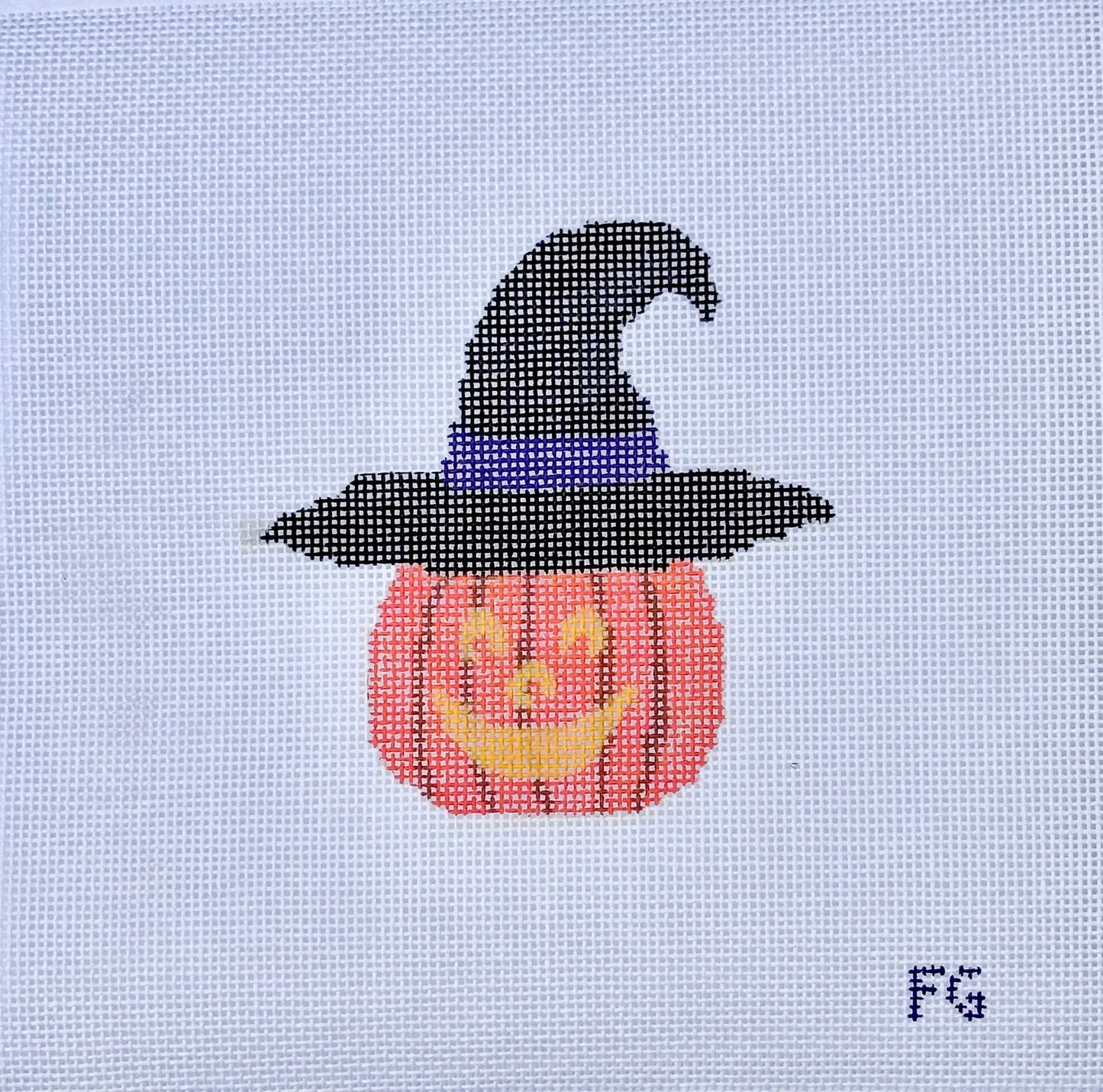 A needlepoint canvas featuring a pixelated design of a jack-o'-lantern pumpkin wearing a black witch hat with a purple band, on a white mesh grid. The initials 'FG' are printed in the bottom right corner.