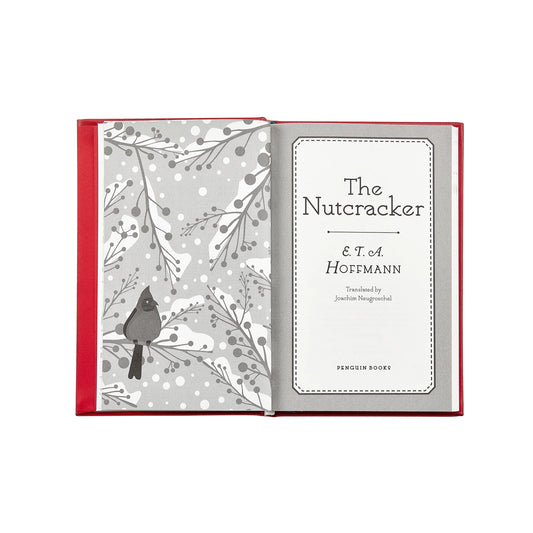 An open hardcover book with a red cover and gray-and-white illustrated interior pages. The left page shows a bird perched among winter branches, and the right page displays the title 'The Nutcracker & T.