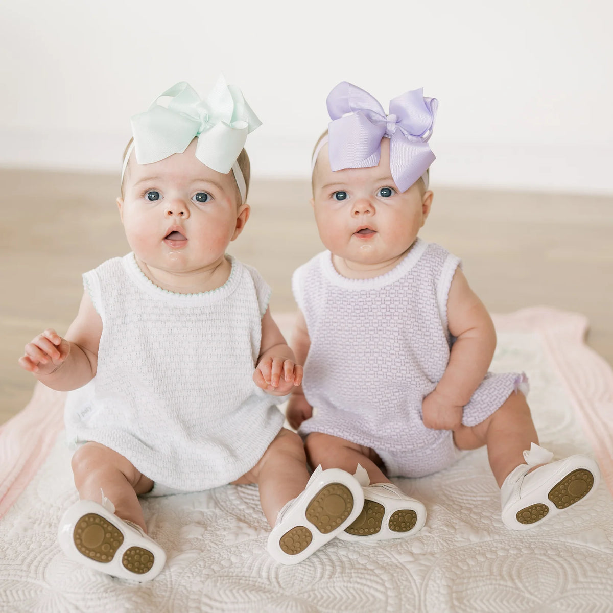 Two babies sitting side by side wearing matching textured sleeveless outfits and large bow headbands, one in white and one in pale lavender, photographed in a soft neutral indoor setting.