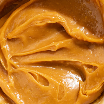 A close-up texture shot of smooth, glossy caramel-colored peanut butter with visible swirls and creamy highlights.