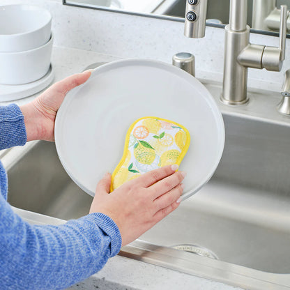 Person using a yellow sponge with a lemon design to clean a white plate in a kitchen sink.