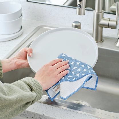 Person cleaning a white plate with a blue patterned microfiber cloth in a kitchen.