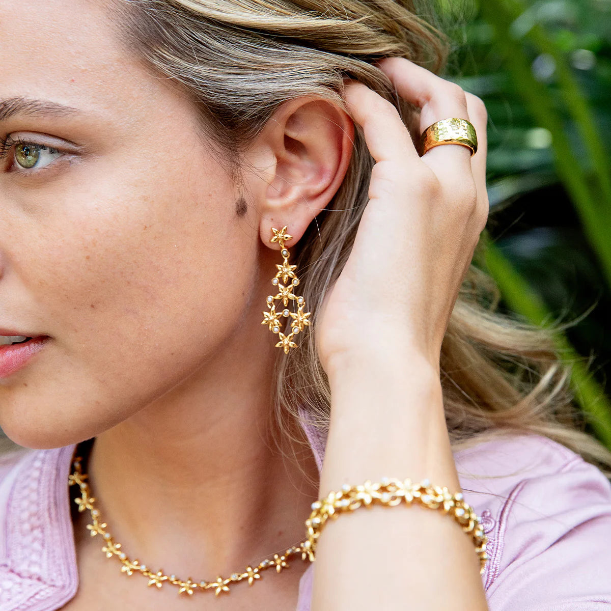 Woman wearing gold jewelry including earrings, necklace, and bracelet with a blurred green background