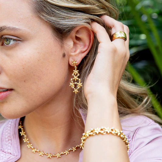 Woman wearing gold jewelry including earrings, necklace, and bracelet with a blurred green background