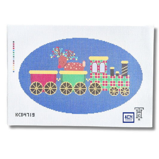 A needlepoint canvas featuring a colorful Christmas train design. The train has pink, green, and plaid red-and-white carriages with gold wheels, set against a blue oval on a white canvas background.