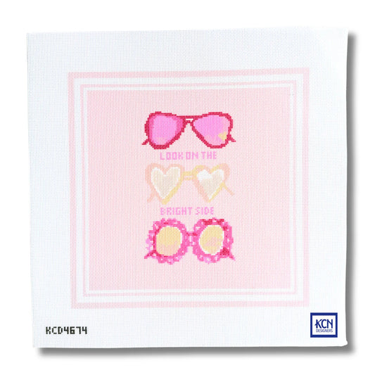 A square needlepoint canvas featuring a pink and white graphic design with three pairs of heart-shaped sunglasses and the text 'LOOK ON THE BRIGHT SIDE'. The canvas shows the KCN Designs brand logo in the bottom right corner.