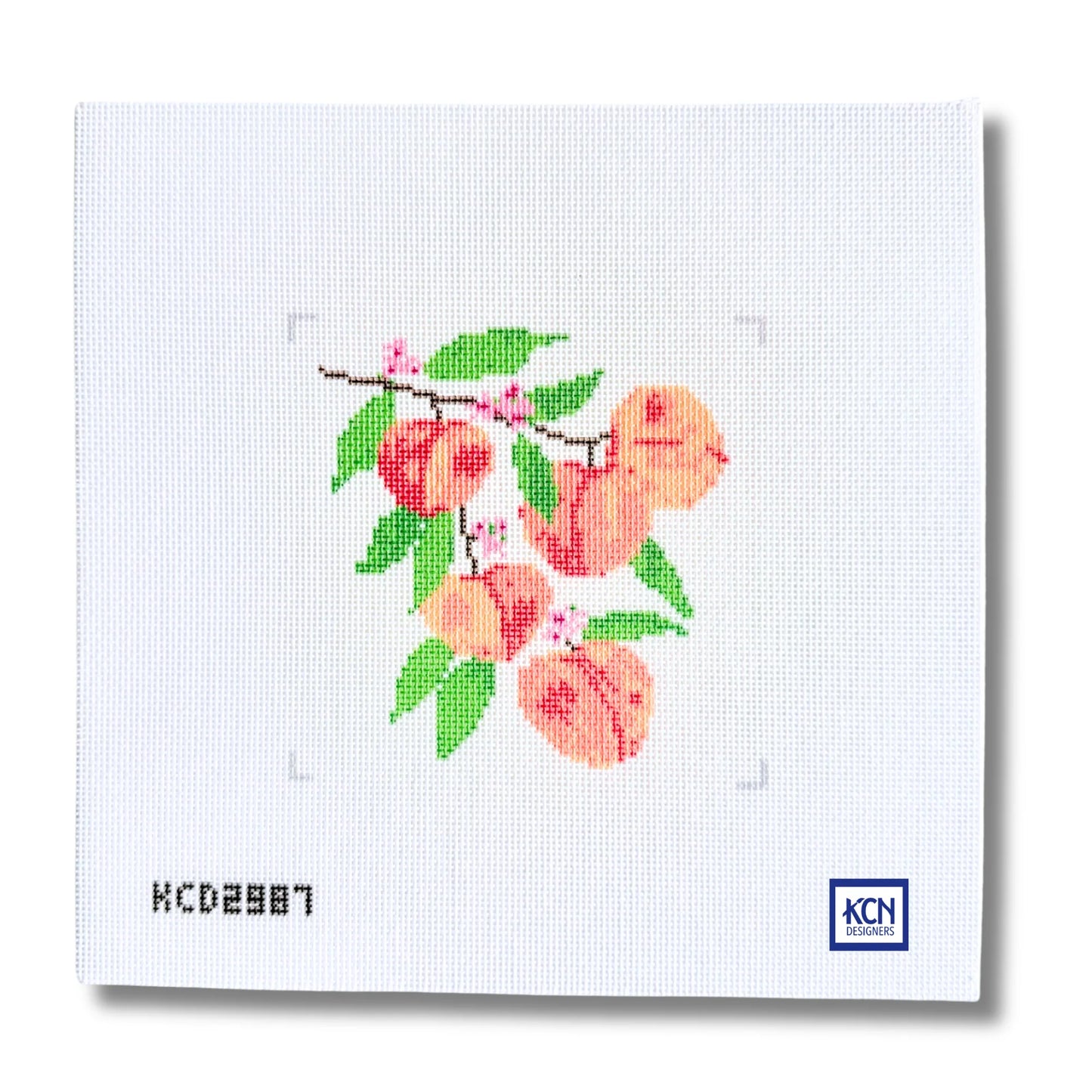 A square white needlepoint canvas featuring a stitched peach branch with green leaves and pink blossoms, shown on a clean white background with a small KCN logo in the bottom right corner.