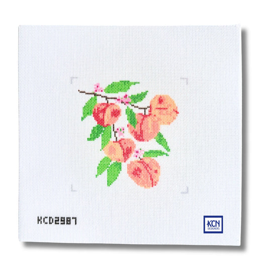 A square white needlepoint canvas featuring a stitched peach branch with green leaves and pink blossoms, shown on a clean white background with a small KCN logo in the bottom right corner.