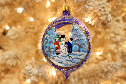 A round blue Christmas ornament with a gold-trimmed border and a hand-painted winter scene featuring children and a snowman, hanging on a softly blurred white Christmas tree with warm lights.