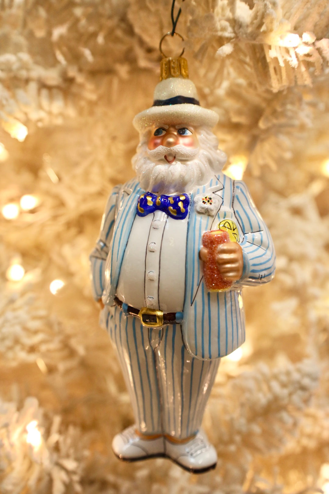A festive Christmas ornament shaped like a jovial old man in a blue and white pinstripe suit, complete with a blue patterned bow tie, white hat, and beard, holding a drink and set against a warm, glowing Christmas tree background.
