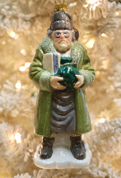 A ceramic Christmas ornament depicting a stern, grumpy old man in a green coat holding a green sack, set against a blurred, warm-lit Christmas tree background.