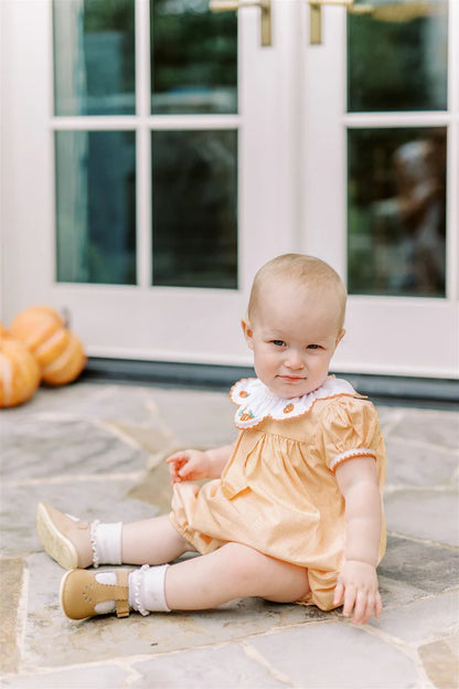 Baby in an orange dress sitting on a stone patio with pumpkins in the background