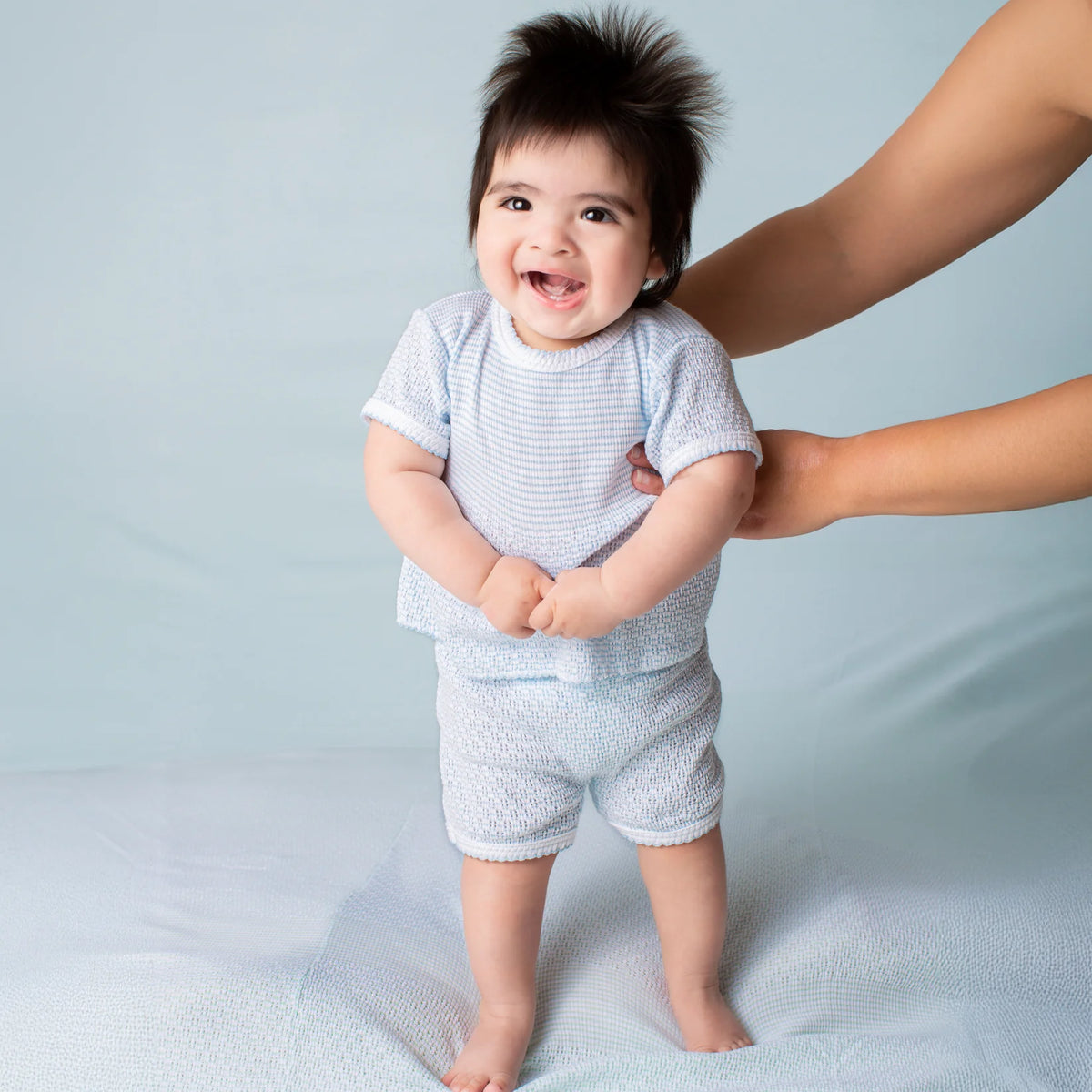 A baby wearing a light blue and white textured short-sleeve shirt and matching shorts stands barefoot on a light blue studio floor while being supported by an adult's hands.