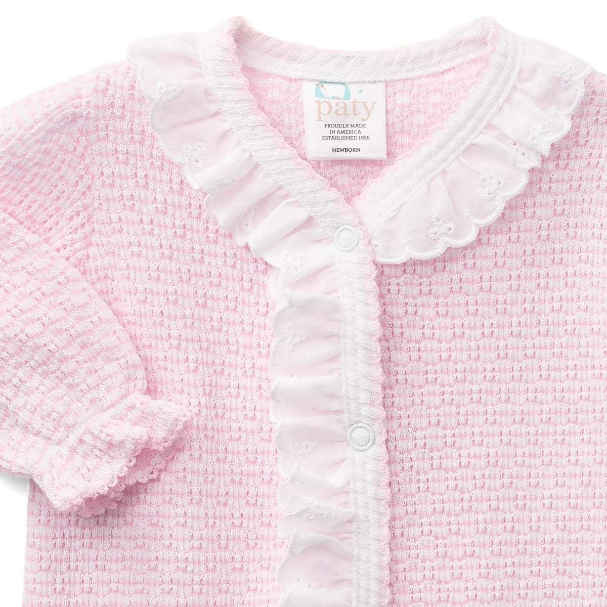 A close-up of a baby cardigan in light pink and white. The cardigan features a textured waffle knit body, a white ruffled collar, a ruffled button placket, and a ruffled sleeve cuff.