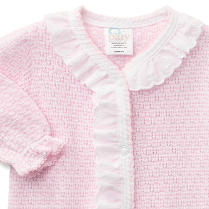A close-up of a baby cardigan in light pink and white. The cardigan features a textured waffle knit body, a white ruffled collar, a ruffled button placket, and a ruffled sleeve cuff.