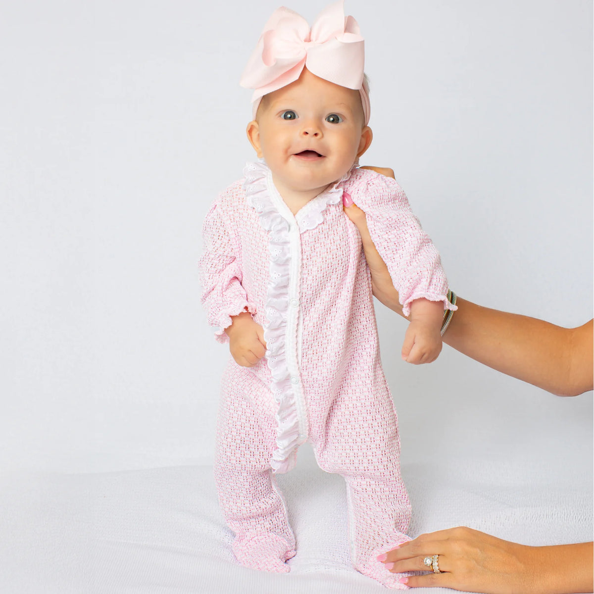 A baby wearing a pink textured footed sleeper with white ruffle trim and a matching pink bow headband, being supported by an adult's hands against a light gray studio background.