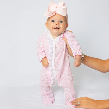 A baby wearing a pink textured footed sleeper with white ruffle trim and a matching pink bow headband, being supported by an adult's hands against a light gray studio background.