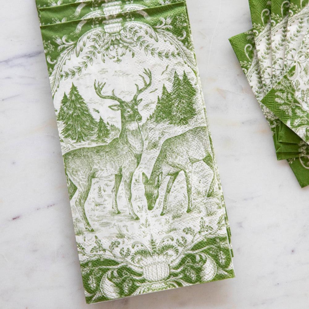 A green and white patterned paper napkin featuring a deer and forest design, laid flat on a light marble surface with a stack of matching napkins partially visible on the right.