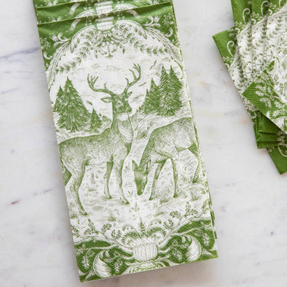 A green and white patterned paper napkin featuring a deer and forest design, laid flat on a light marble surface with a stack of matching napkins partially visible on the right.