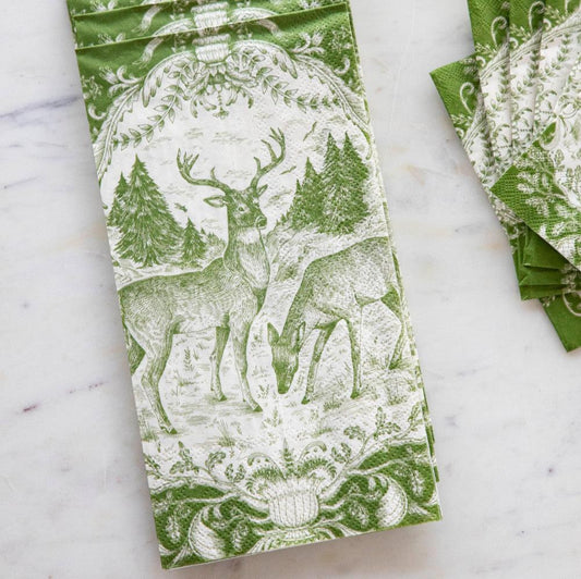A green and white patterned paper napkin featuring a deer and forest design, laid flat on a light marble surface with a stack of matching napkins partially visible on the right.