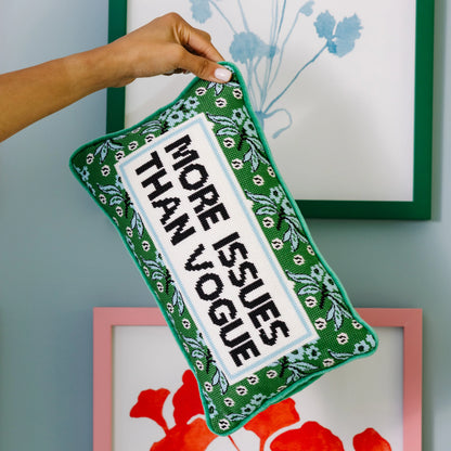 Decorative pillow with text 'More Issues Than Vogue' held by a hand against a wall with framed artwork.