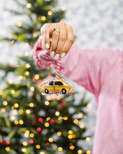 Hand holding a Christmas ornament shaped like a taxi in front of a decorated tree.