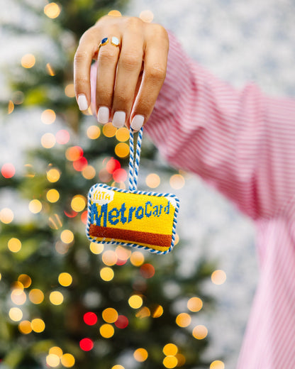 Hand holding a 'Little MetroCard' toy with a blurred Christmas tree in the background