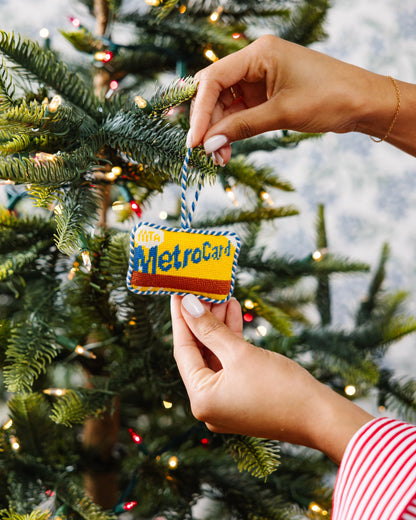 Hand holding a small 'Little MetroCard' ornament against a Christmas tree background