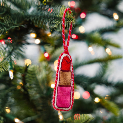 Decorative ornament shaped like a lipstick hanging on a Christmas tree with lights in the background.