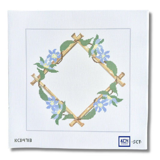 A needlepoint canvas featuring a diamond-shaped wreath design made of tan branches with green leaves and blue flowers, on a white canvas background with printed codes and a KCN logo.