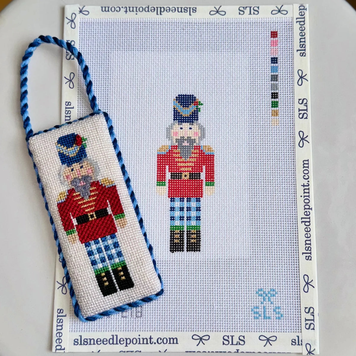 A needlepoint canvas and matching pouch featuring a nutcracker design, displayed on a white branded SLS needlepoint mat. The pouch has a blue braided strap and the canvas shows the stitched design alongside the unworked border and color swatches.