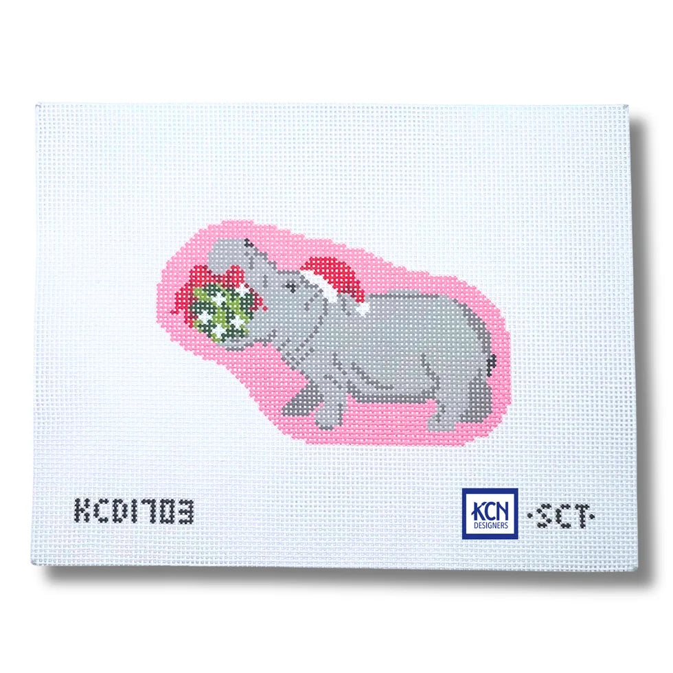 A square white needlepoint canvas featuring a pixelated gray hippopotamus wearing a red Santa hat and holding a green gift with white dots, set against a pink shape. The canvas has a printed serial number 'KCD1703' on the bottom left and a 'KCN Needlework' logo on the bottom right.