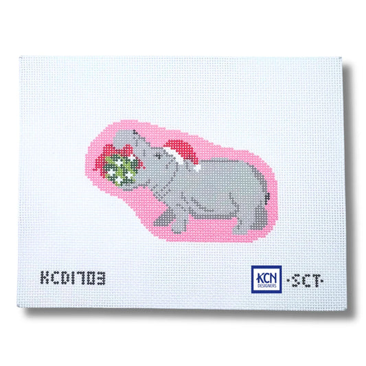 A square white needlepoint canvas featuring a pixelated gray hippopotamus wearing a red Santa hat and holding a green gift with white dots, set against a pink shape. The canvas has a printed serial number 'KCD1703' on the bottom left and a 'KCN Needlework' logo on the bottom right.