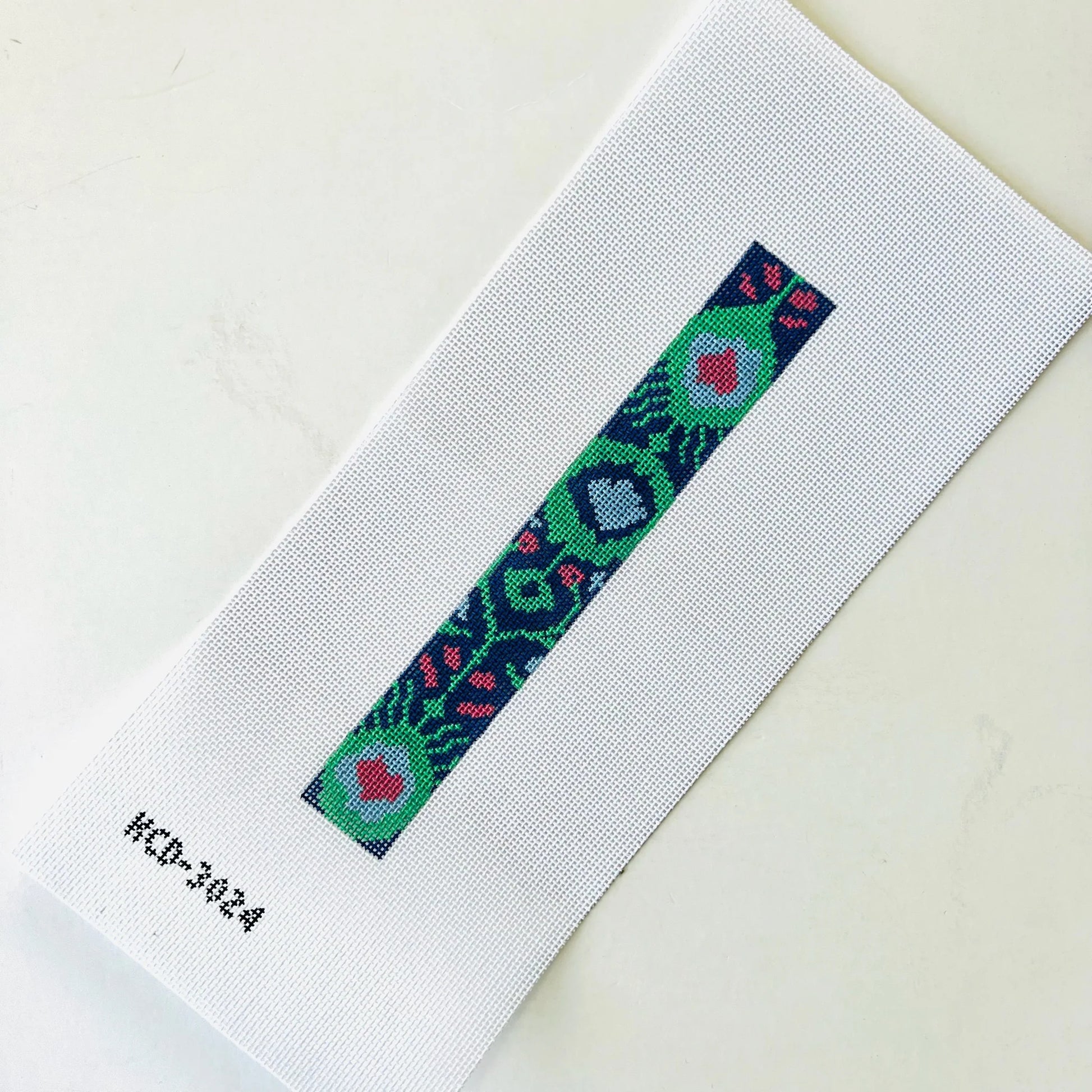 A close-up of a white woven fabric strap or band with a narrow central embroidered geometric pattern in green, blue, pink, and red, photographed on a light neutral background. Small printed text 'HCD-3024' appears near the lower left.