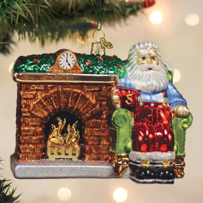 Christmas ornament of Santa Claus by a fireplace with a clock above, on a blurred Christmas tree background.