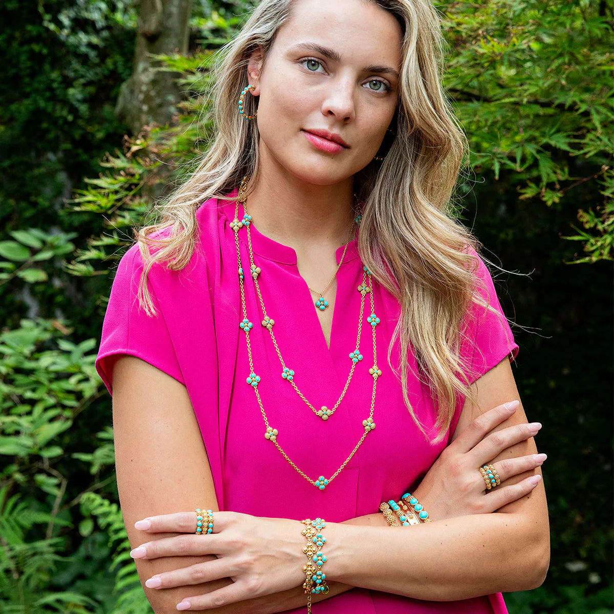 Woman wearing a bright pink top with layered necklaces and bracelets against a green foliage background