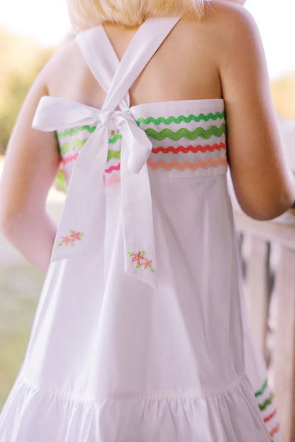 A white Sundress with multicolored ric rac trim and a bow at the back neck, designed for children.