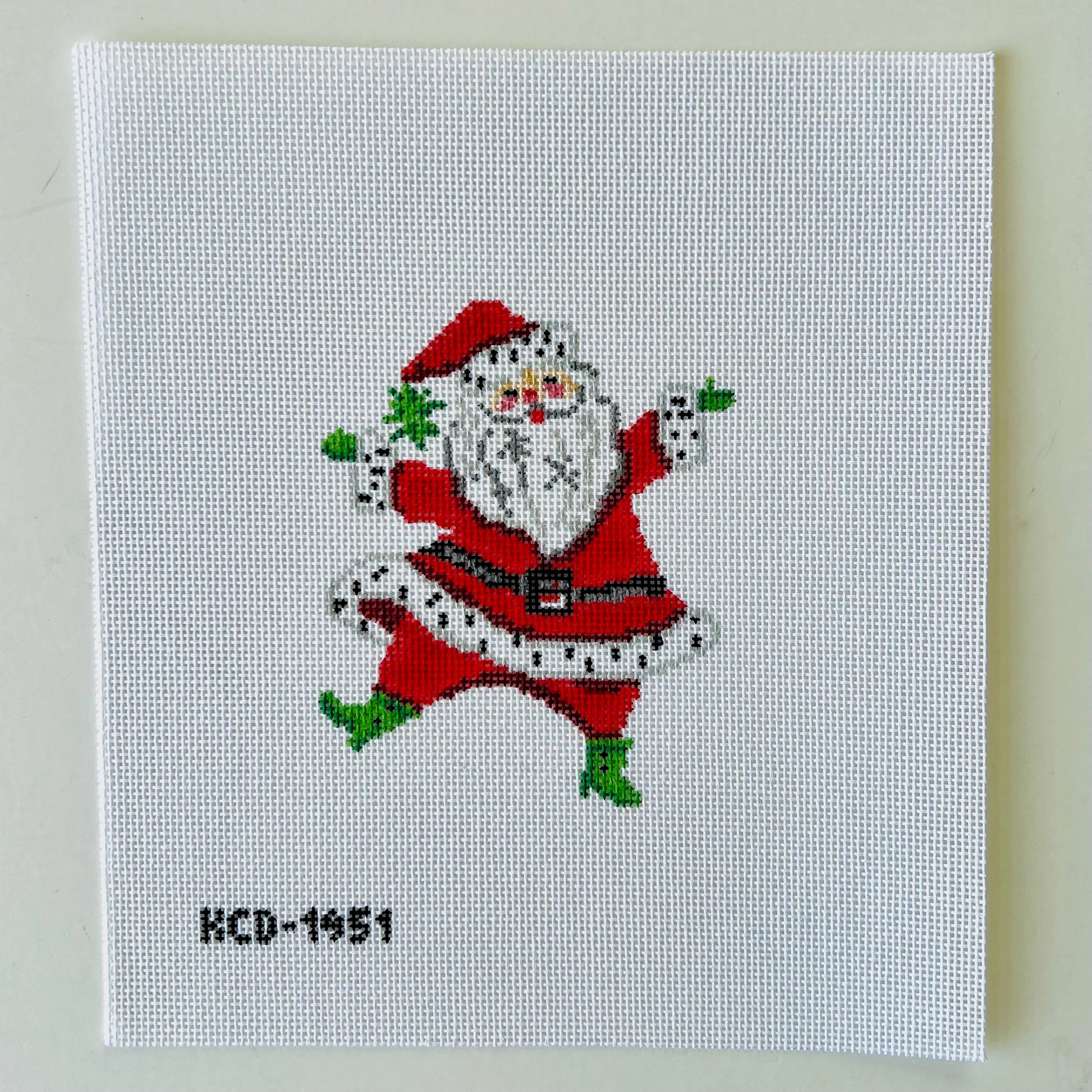 A square white needlepoint canvas featuring a colorful embroidered-style Santa Claus figure in the center and the text 'KCD-1951' in the lower left corner, photographed against a light neutral background.