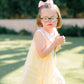 Young girl in a yellow dress with a pink bow standing outdoors on grass.