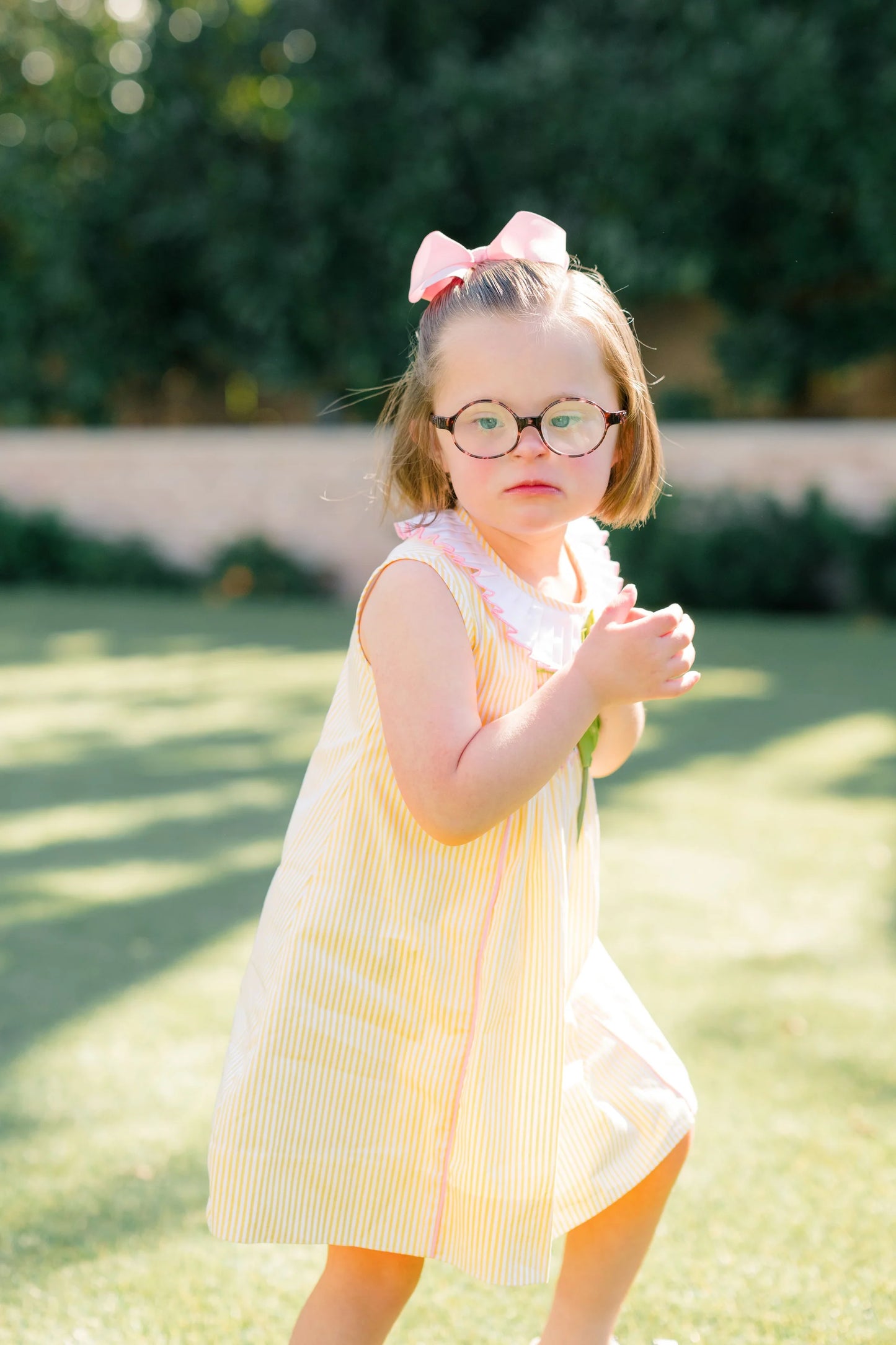 Young girl in a yellow dress with a pink bow standing outdoors on grass.
