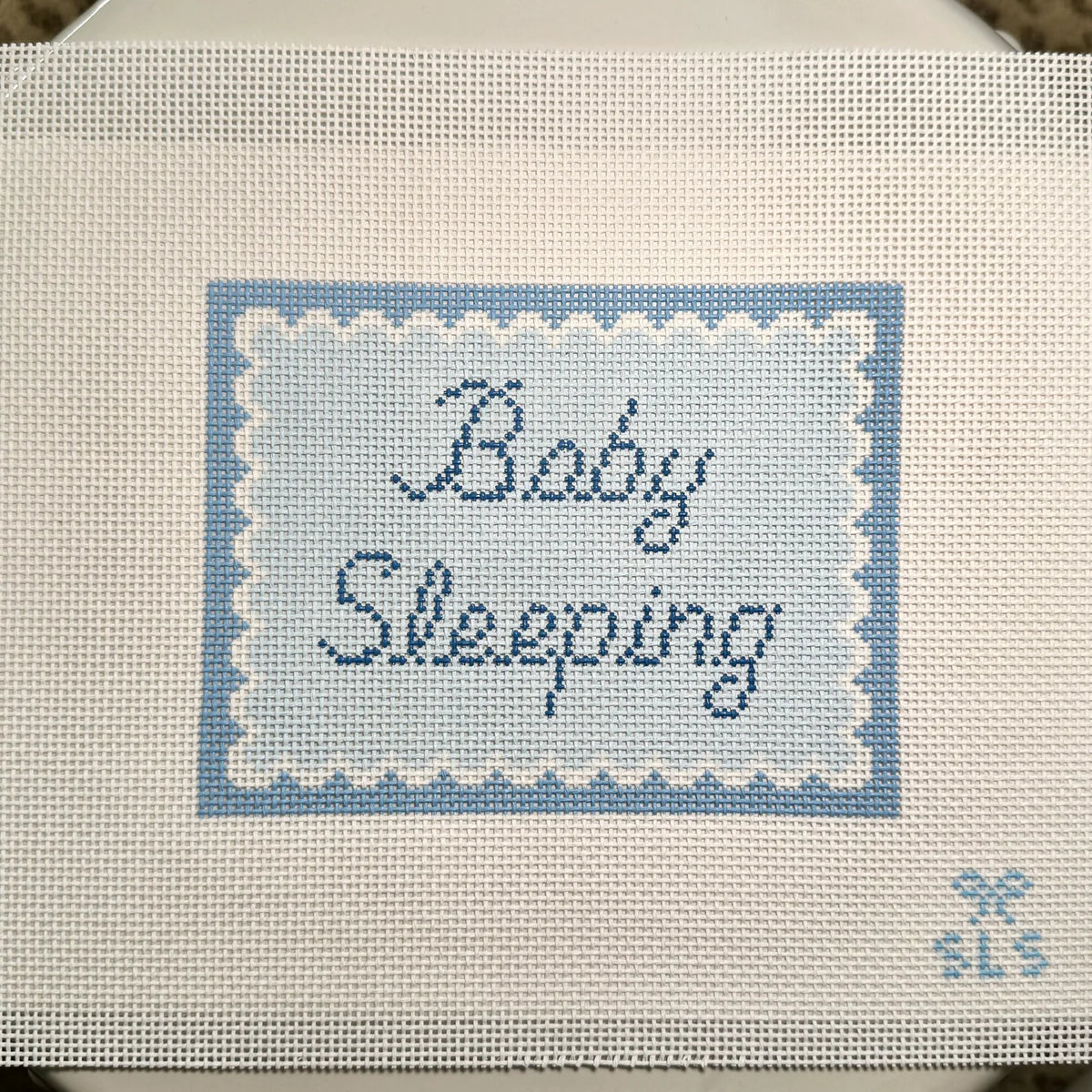 A close-up of a cross-stitch canvas featuring the words 'Baby Sleeping' stitched in navy blue script inside a light blue and white scalloped border. The canvas is made of white Aida fabric with a small 'SLS' monogram in the bottom right corner.