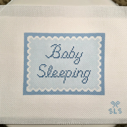 A close-up of a cross-stitch canvas featuring the words 'Baby Sleeping' stitched in navy blue script inside a light blue and white scalloped border. The canvas is made of white Aida fabric with a small 'SLS' monogram in the bottom right corner.